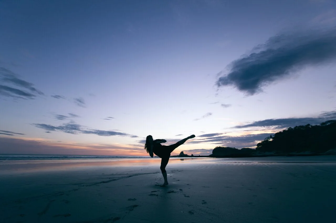 Silhouette of a person performing a high kick on a beach at dusk.