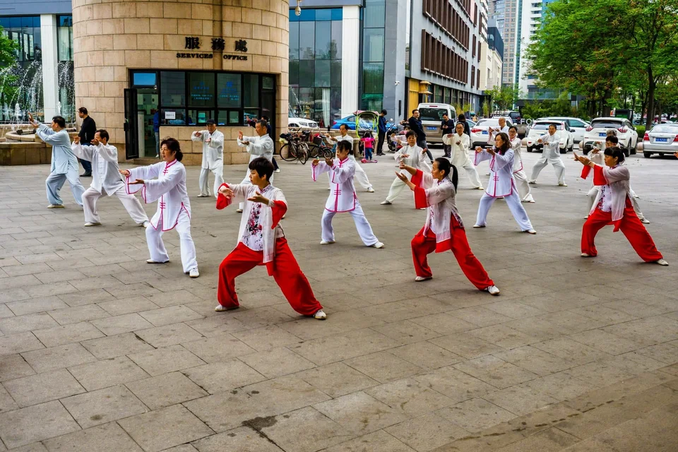 A group practicing Tai Chi in an outdoor plaza, wearing white uniforms with red or pink trim.